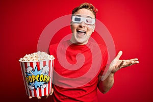 Young handsome redhead man watching 3d glasses eating popcorn snack over red background very happy and excited, winner expression