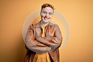 Young handsome redhead man wearing casual leather jacket over isolated yellow background happy face smiling with crossed arms