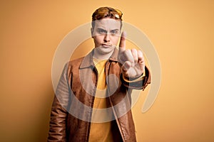 Young handsome redhead man wearing casual leather jacket over isolated yellow background Pointing with finger up and angry