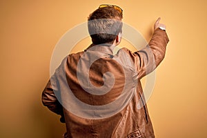 Young handsome redhead man wearing casual leather jacket over isolated yellow background Posing backwards pointing ahead with