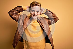 Young handsome redhead man wearing casual leather jacket over isolated yellow background relaxing and stretching, arms and hands