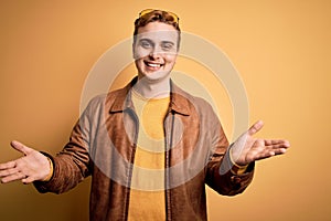 Young handsome redhead man wearing casual leather jacket over isolated yellow background smiling cheerful offering hands giving