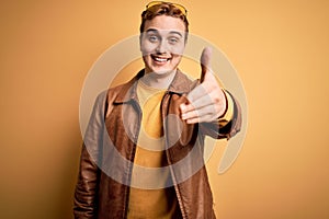 Young handsome redhead man wearing casual leather jacket over isolated yellow background smiling friendly offering handshake as