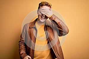 Young handsome redhead man wearing casual leather jacket over isolated yellow background smiling and laughing with hand on face