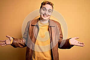 Young handsome redhead man wearing casual leather jacket over isolated yellow background smiling showing both hands open palms,