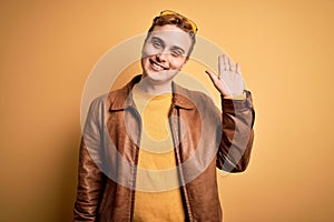 Young handsome redhead man wearing casual leather jacket over isolated yellow background Waiving saying hello happy and smiling,