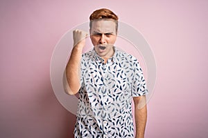 Young handsome redhead man wearing casual summer shirt standing over pink background angry and mad raising fist frustrated and