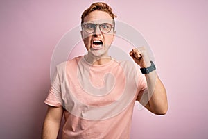 Young handsome redhead man wearing casual t-shirt standing over isolated pink background angry and mad raising fist frustrated and