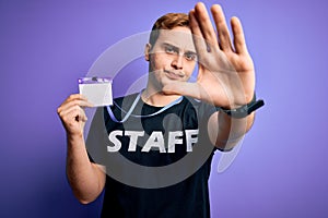 Young handsome redhead worker man wearing staff t-shirt uniform holding id card pass with open hand doing stop sign with serious