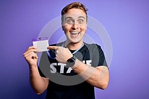 Young handsome redhead worker man wearing staff t-shirt uniform holding id card pass Smiling happy pointing with hand and finger