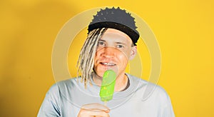 Young handsome satisfied man with dreadlocks in black cap eating ice cream on yellow background. Joyful guy in blue t