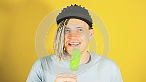 Young handsome satisfied man with dreadlocks in black cap eating ice cream on yellow background. Joyful guy in blue t