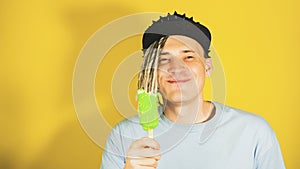 Young handsome satisfied man with dreadlocks in black cap eating ice cream on yellow background. Joyful guy in blue t