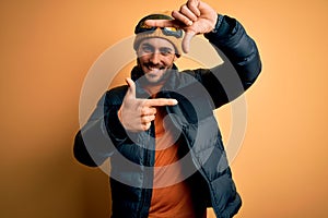 Young handsome skier man with beard wearing snow sportswear and ski goggles smiling making frame with hands and fingers with happy