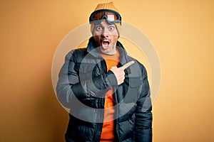 Young handsome skier man skiing wearing snow sportswear using ski goggles Surprised pointing with finger to the side, open mouth