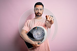 Young handsome slim sportsman with beard holding scale over isolated pink background pointing with finger to the camera and to