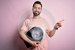 Young handsome slim sportsman with beard holding scale over isolated pink background very happy pointing with hand and finger to