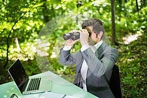 Young handsome smile business man at work table office with laptop in green forest with binocular looking for competitors. Busines