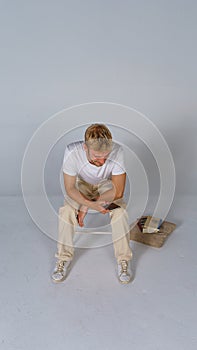 Young handsome smiling blond man wearing casual t-shirt and beige pants on white background and sitting with curved back looking