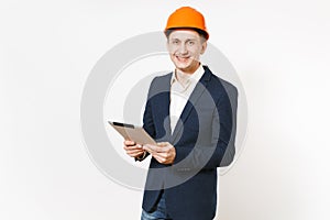 Young handsome smiling businessman in dark suit, protective construction helmet holding tablet pc computer isolated on