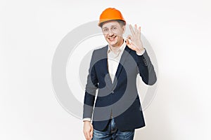 Young handsome smiling businessman in dark suit, protective construction orange helmet showing OK gesture isolated on