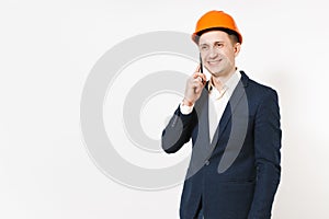 Young handsome smiling businessman in dark suit, protective construction orange helmet talking on mobile phone isolated