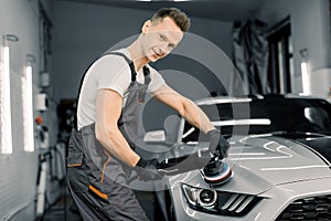 Young handsome smiling Caucasian man, worker of auto detailing service, holds a polisher in the hand and polishes the