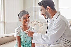 Young handsome smiling doctor using a stethoscope to listen to a sick little girls heartbeat during a house call checkup
