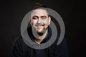 A young handsome smiling man in a blue hoodie is sitting and looking at the camera. Black background. Close-up