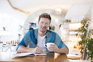 Young handsome smiling man drinks coffee during his lunch break in the cafe taking notes to his planner looking busy, multitasking