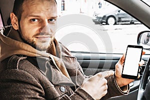 Young handsome smiling man showing smartphone or cellphone white screen as mock up for your product sitting in car