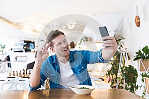 Young handsome smiling man speaking online in his smartphone while eating salad during his lunch break in the cafe , multitasking