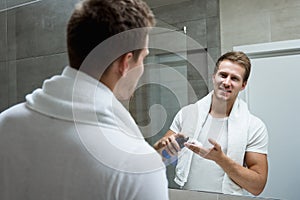 Young handsome smiling man with white towel on his shoulders looking in the bathroom mirror putting shaving foam on his