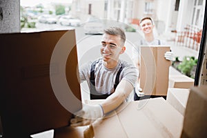 A young handsome smiling mover wearing uniform is reaching for the box while unloading the van full of boxes. House move