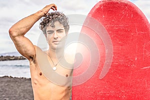 Young handsome spanish man with curly hair standing shirtless on the beach with red surfboard, smiling to the camera. Good vibes