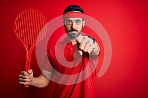 Young handsome sportsman with beard playing tennis using racket over red background pointing with finger to the camera and to you,
