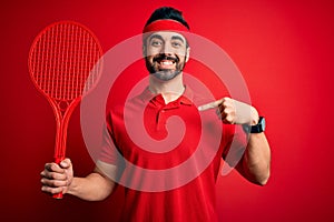 Young handsome sportsman with beard playing tennis using racket over red background with surprise face pointing finger to himself