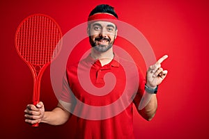 Young handsome sportsman with beard playing tennis using racket over red background very happy pointing with hand and finger to