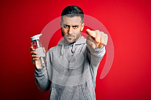 Young handsome sportsman drinking bottle of water to refeshment over red background pointing with finger to the camera and to you,