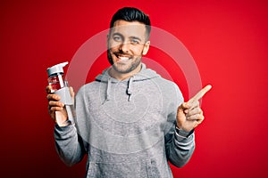 Young handsome sportsman drinking bottle of water to refeshment over red background very happy pointing with hand and finger to