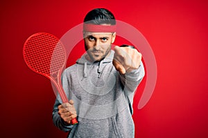Young handsome sportsman holding tennis racket wearing sportswear over red background pointing with finger to the camera and to