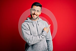 Young handsome sportsman wearing sweatshirt standing over isolated red background cheerful with a smile of face pointing with hand