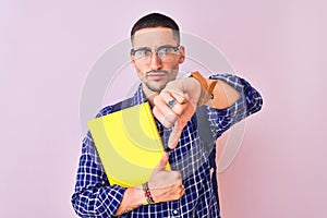 Young handsome student man holding a book over isolated background with angry face, negative sign showing dislike with thumbs