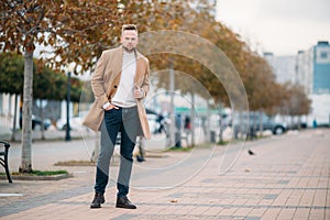 Young handsome stylish man wiyh beard in elegant coat standing in autumn park