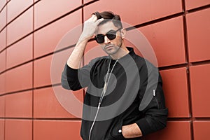 Young handsome stylish model man in sunglasses straightens his hair. Handsome guy in black trendy jacket poses near the metal wall