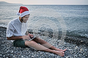 Young handsome successful freelance businessman is sitting on rocky beach in red Santa hat and working on laptop during holidays.