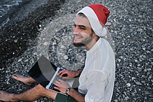 Young handsome successful freelance businessman is sitting on rocky beach in red Santa hat and working on laptop during holidays.