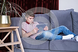 Young handsome teenager boy using tablet sitting on grey sofa at home