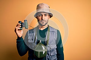 Young handsome tourist man with beard on vacation wearing explorer hat using binoculars with a confident expression on smart face