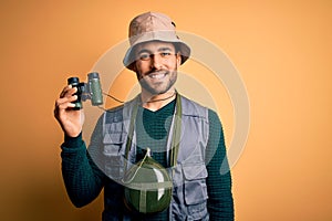 Young handsome tourist man with beard on vacation wearing explorer hat using binoculars with a happy face standing and smiling